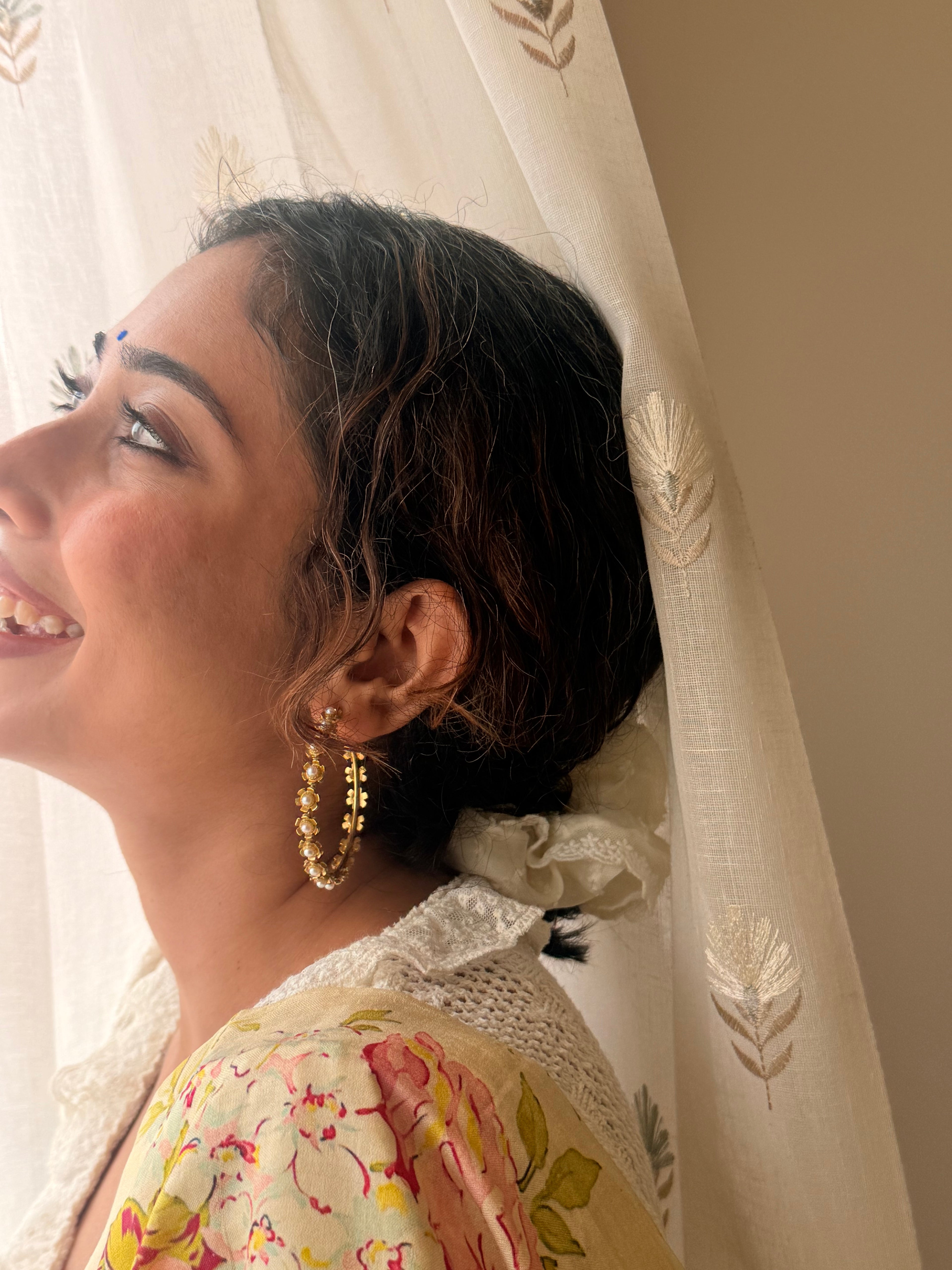 Woman smiling and looking upward, wearing Gulnaar Hoops with pearl-centred flower detail, sheer embroidered curtain in the background.