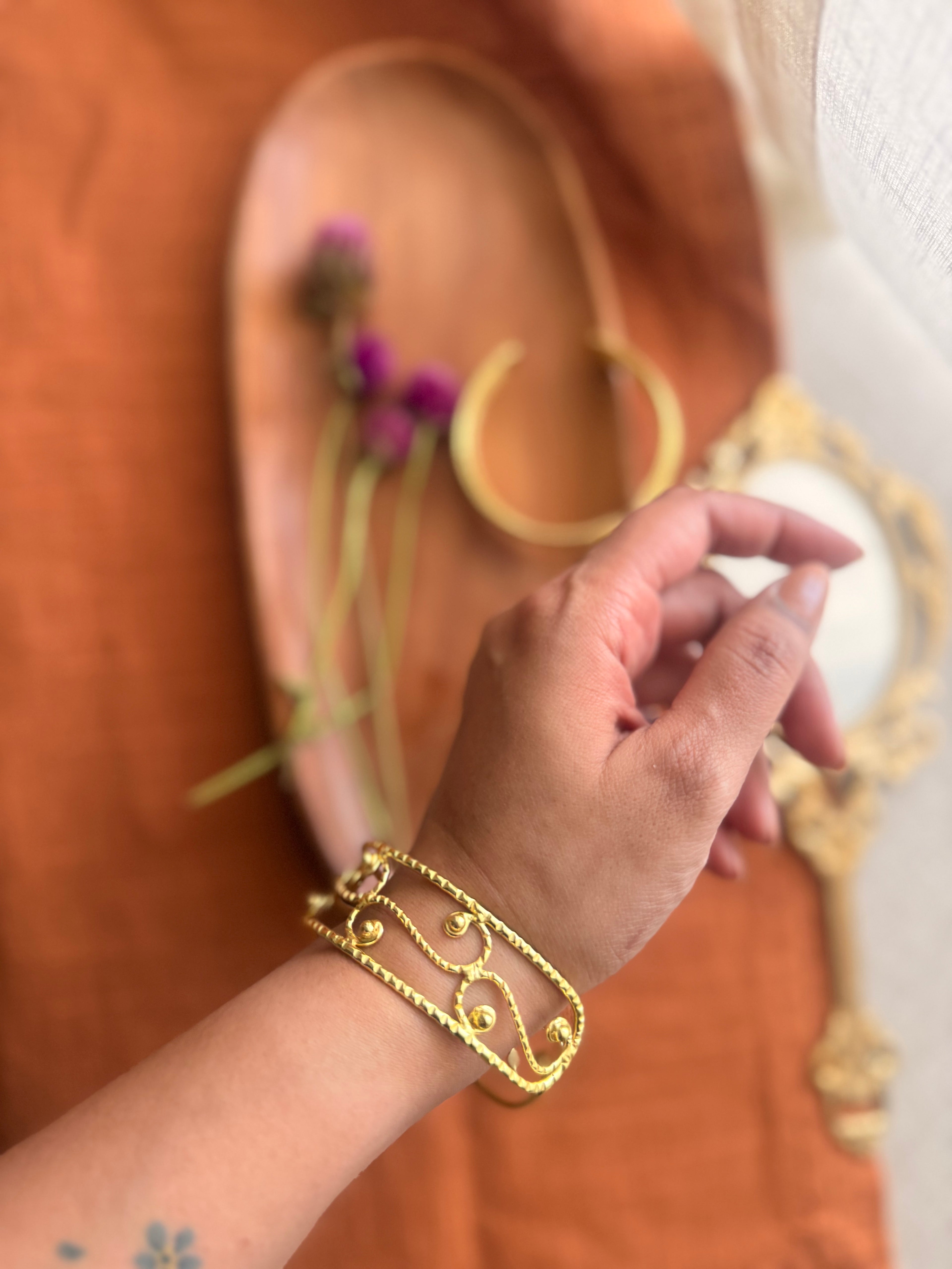 Close-up of a wide gold cuff bracelet with an intricate swirling filigree design and textured edges worn on a person's wrist.