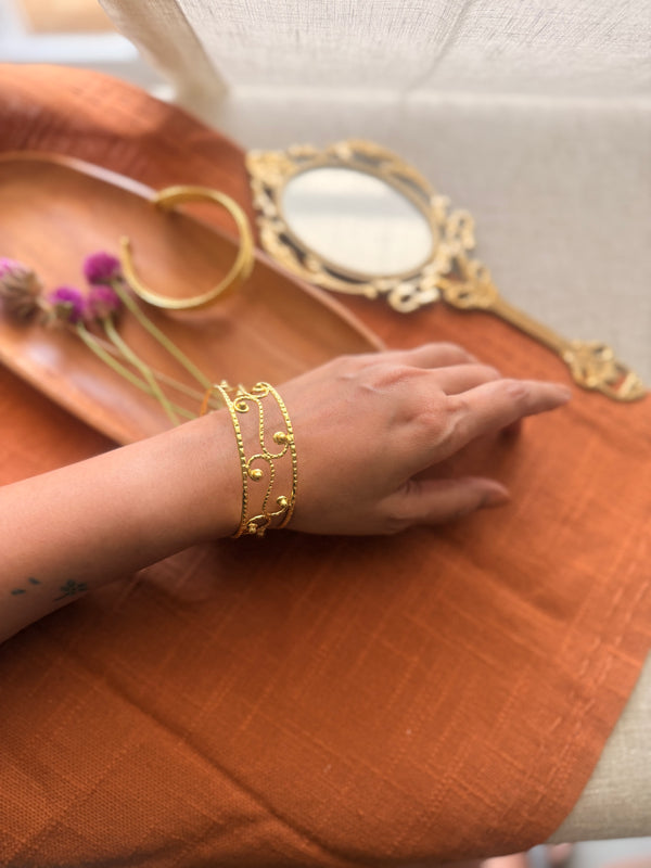 A gold filigree cuff bracelet with open-work scroll patterns and gold bead accents worn on a wrist, set against a warm, sunlit background.