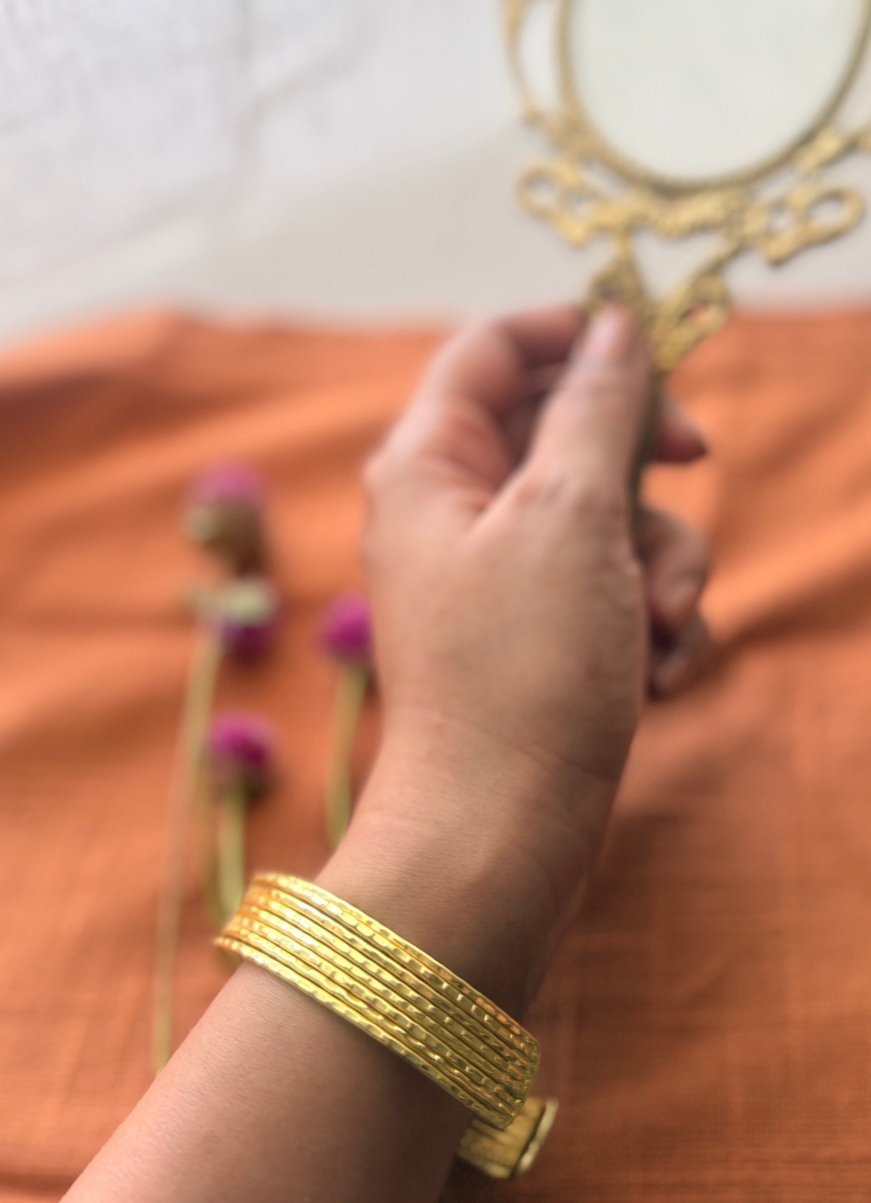 Close-up of a textured gold ribbed cuff bracelet on a wrist, with a soft-focus background featuring a vintage mirror and purple globe amaranth flowers.