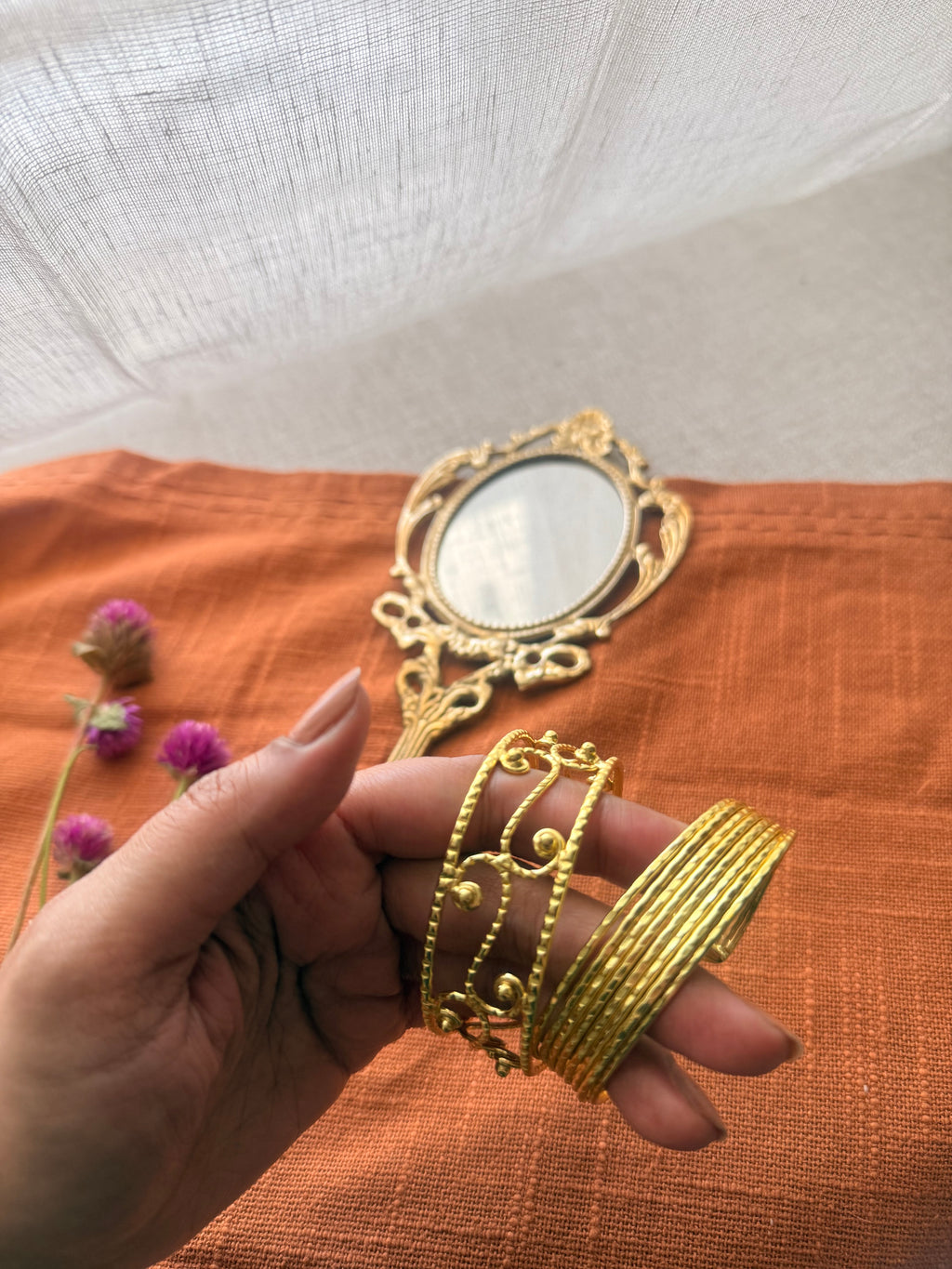 A hand holding two gold cuff bracelets—one with a delicate filigree scroll design and the other with a bold ribbed texture—to show how they can be styled together.
