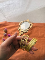 A hand holding two gold cuff bracelets—one with a delicate filigree scroll design and the other with a bold ribbed texture—to show how they can be styled together.