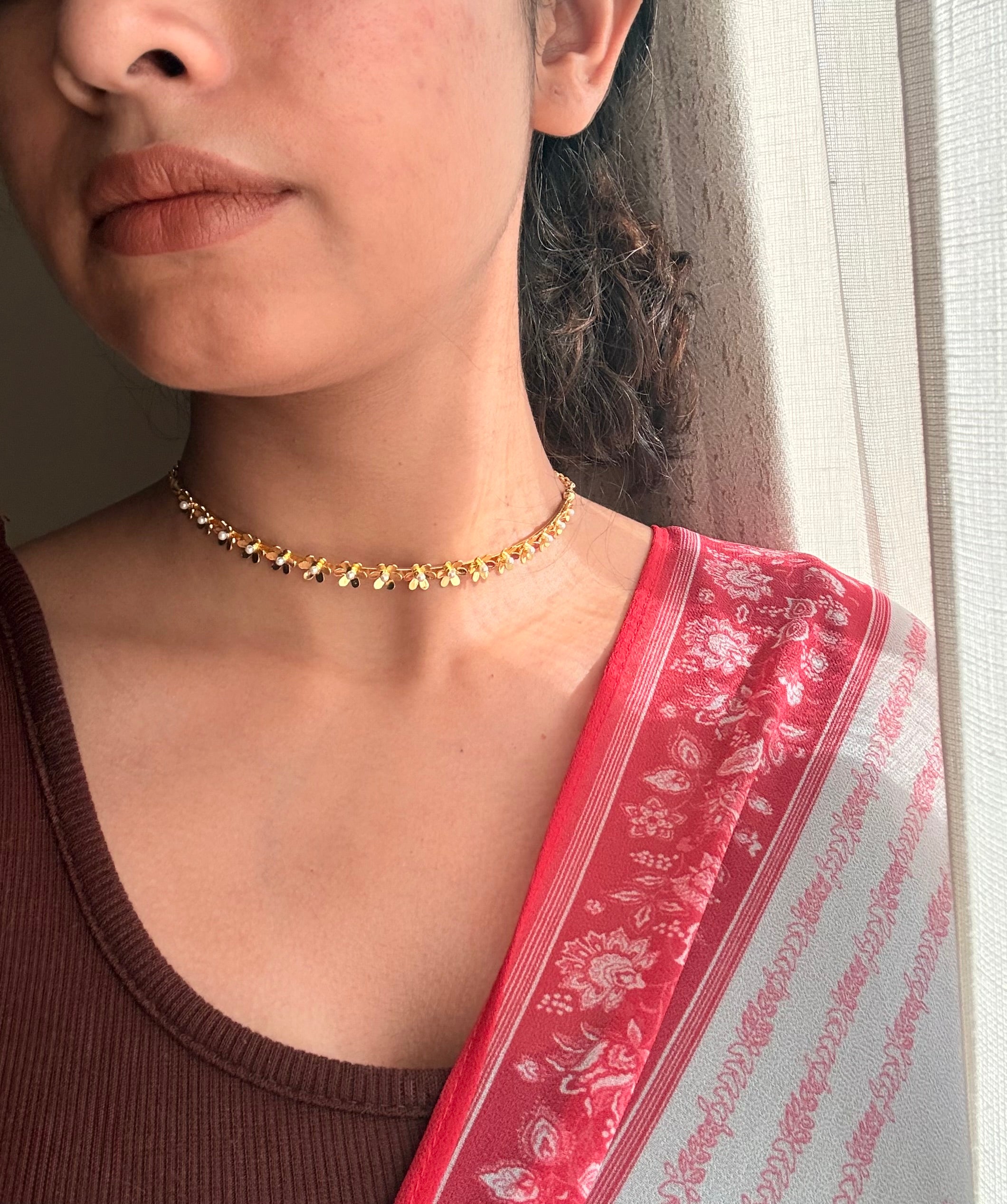 Gold plated brass Motiya Choker with jasmine-shaped flowers worn close to the collarbone, styled with a brown fitted top and a red floral saree dupatta in soft natural window light.