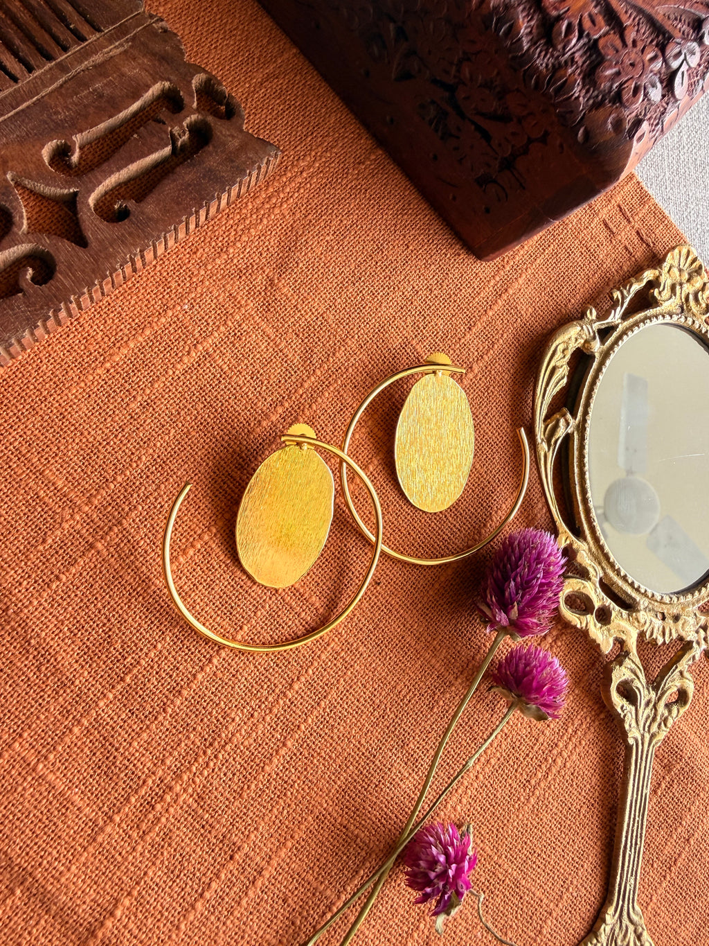 Gold-plated brass hoop earrings with oval textured disc on terracotta fabric, styled with a vintage mirror and dried purple Globe Amaranth flowers