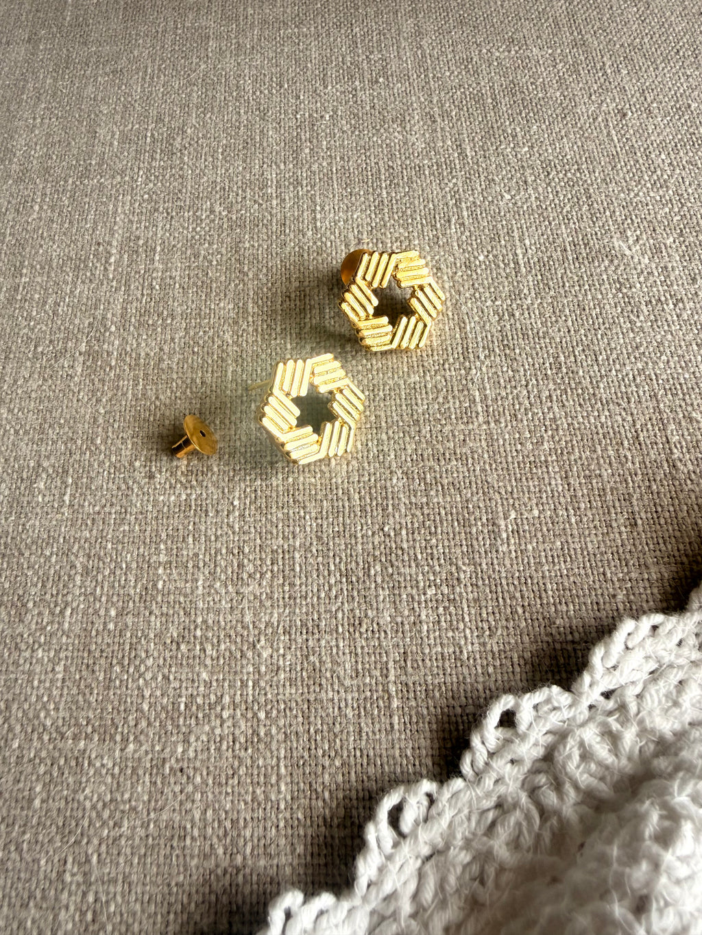 Yantra studs flatlay on grey linen fabric with white lace, showing the hollow hexagon shape and ridged edges, soft natural light