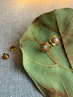 Champa studs resting on a large dried green leaf on grey linen fabric, showing gold five-petal flower design with pearl centre, earring backs placed separately to the left