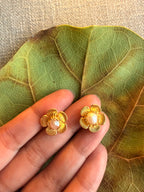Gold flower-shaped earrings with a pearl center held between fingers on a leaf background