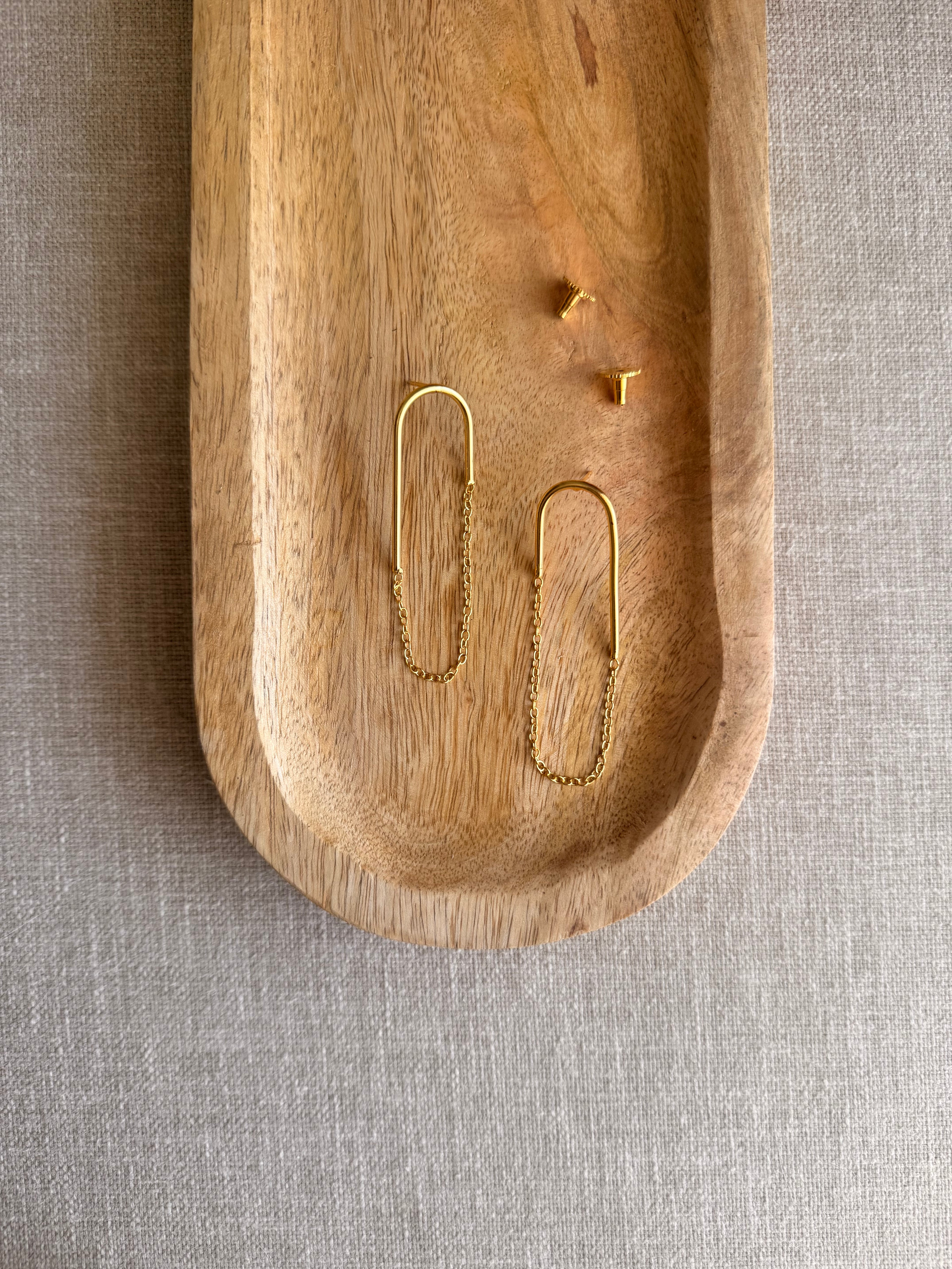 Taar earrings flatlay in a wooden tray, showing the high-low gold wire arc and fine chain detail, earring backs placed separately, grey linen background