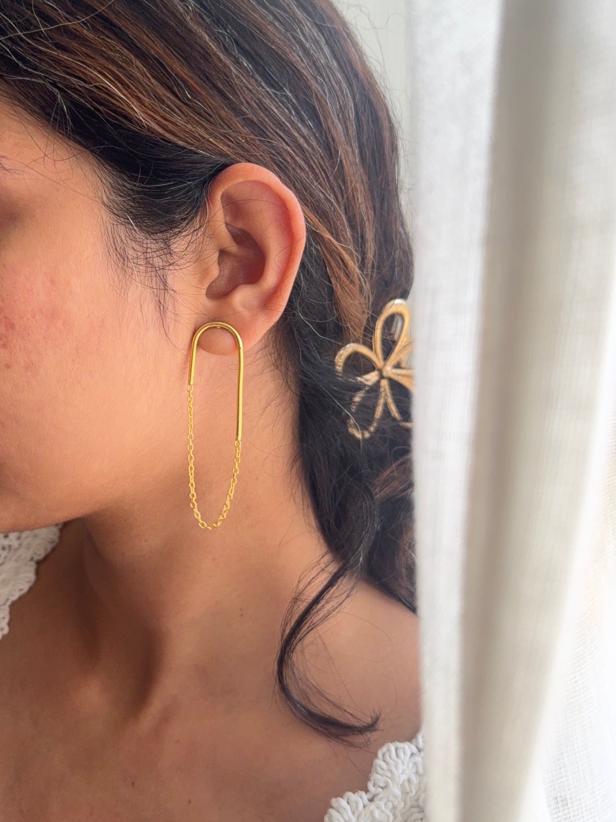 Close-up of woman wearing Taar earrings, gold wire arc and chain visible against her ear, gold flower hair accessory in background, soft natural light through sheer curtain