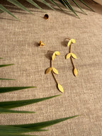 Eafy design Pallavi earrings flatlay on beige linen fabric with palm fronds, showing gold leaf cluster stud and trailing leaf chain detail, earring backs placed separately, soft natural light.
