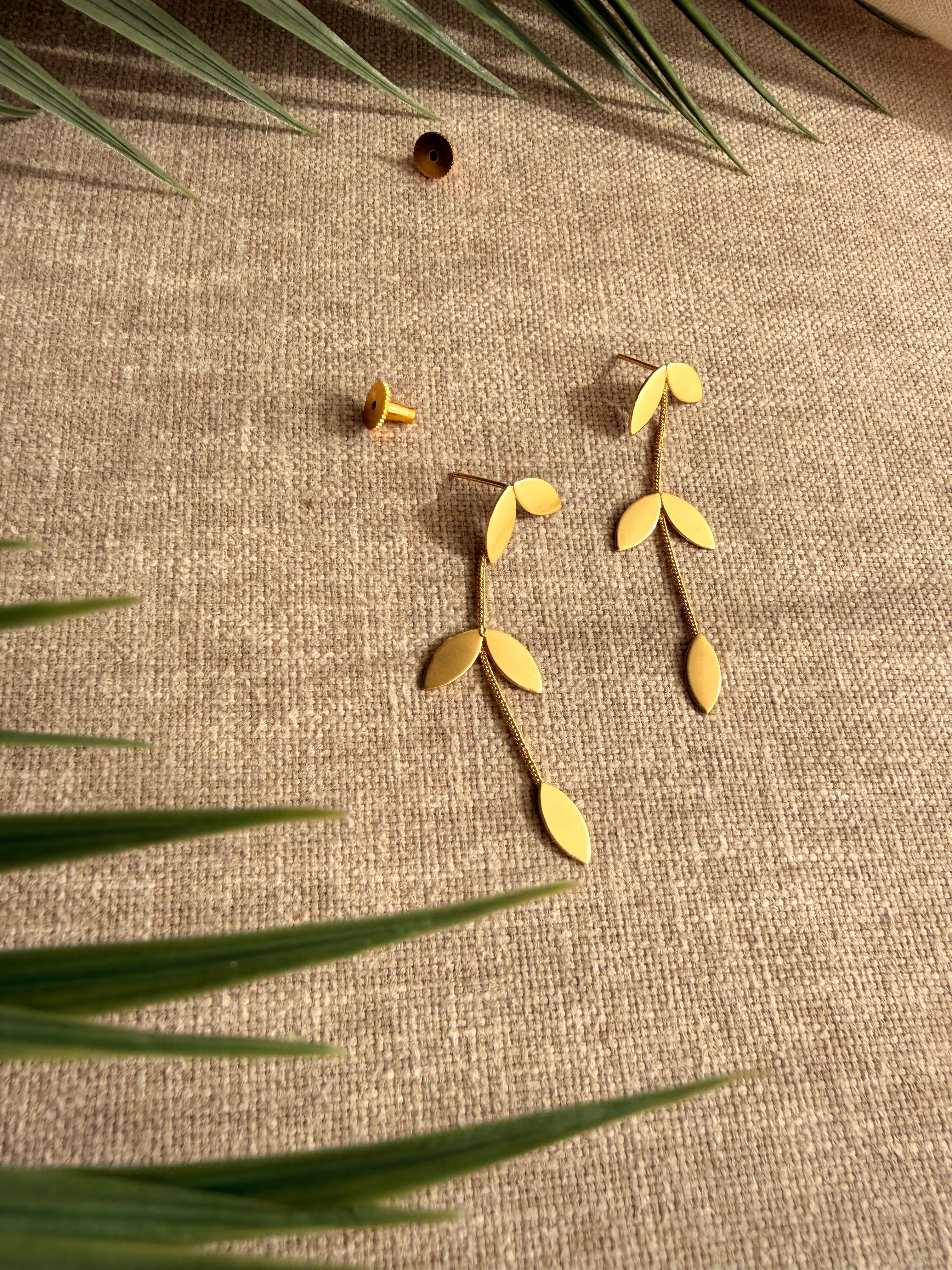 Eafy design Pallavi earrings flatlay on beige linen fabric with palm fronds, showing gold leaf cluster stud and trailing leaf chain detail, earring backs placed separately, soft natural light.