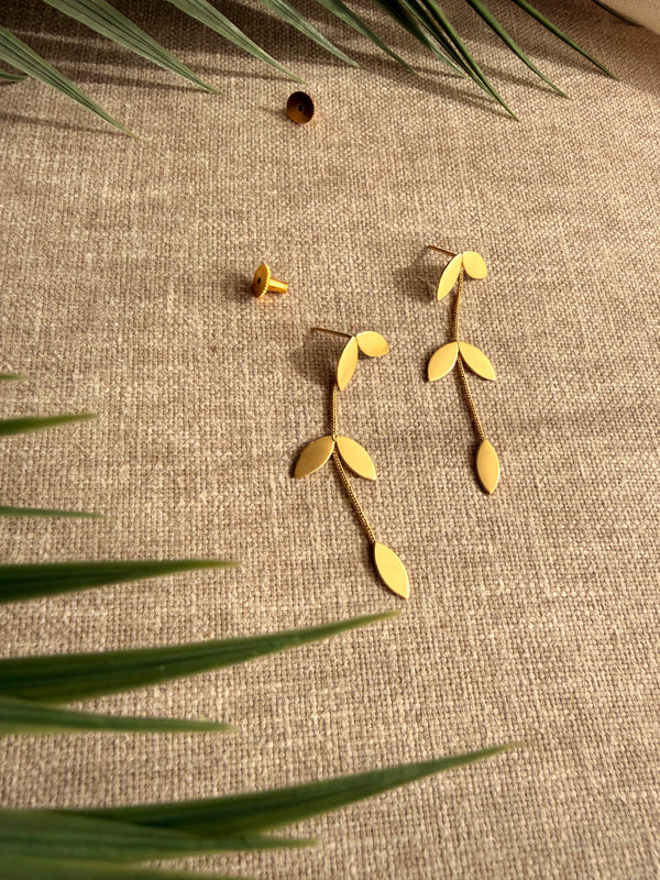 Eafy design Pallavi earrings flatlay on beige linen fabric with palm fronds, showing gold leaf cluster stud and trailing leaf chain detail, earring backs placed separately, soft natural light.