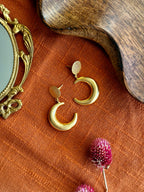 Pair of matte gold Indu Danglers, each a crescent moon hanging from a small textured disc, laid on rust linen beside a brass hand mirror, a wooden tray and dried pink globe amaranth flowers