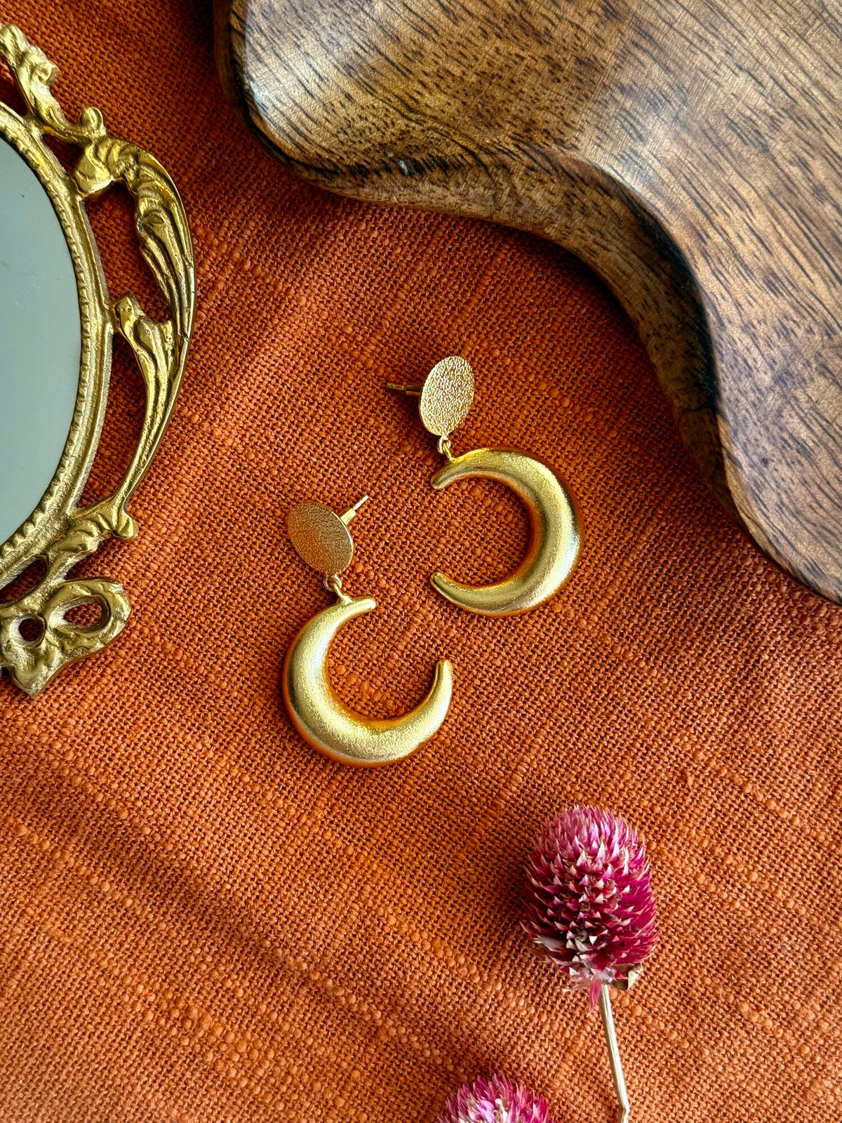 Pair of matte gold Indu Danglers, each a crescent moon hanging from a small textured disc, laid on rust linen beside a brass hand mirror, a wooden tray and dried pink globe amaranth flowers
