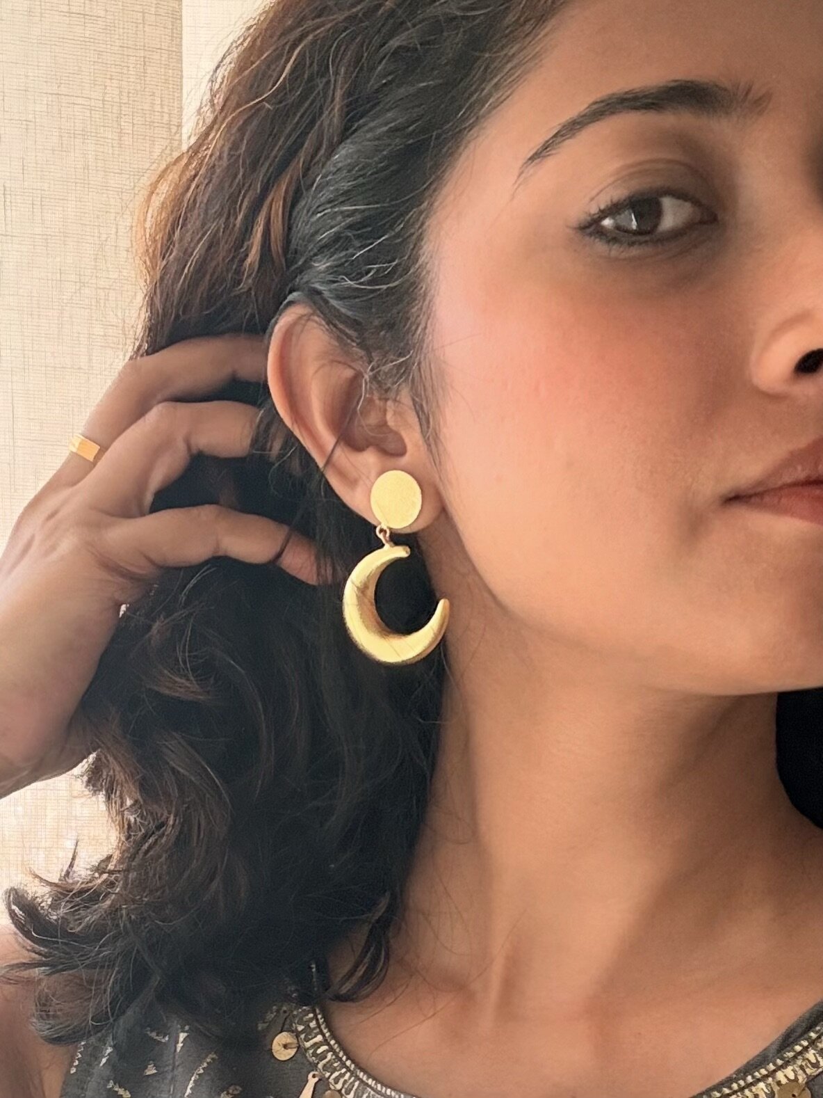 Close-up of a woman tucking her hair behind her ear to show an Indu Dangler, a matte gold crescent hanging from a round textured stud