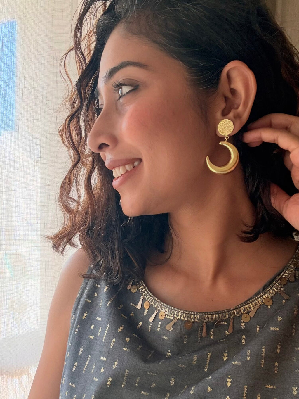 Smiling woman in side profile by a sheer curtain, wearing an Indu Dangler with a gold crescent, in a grey top with a gold sequin and tassel neckline