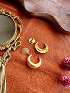 Indu Danglers on rust linen with one earring turned to show its back and post, beside an antique brass hand mirror and dried pink flowers