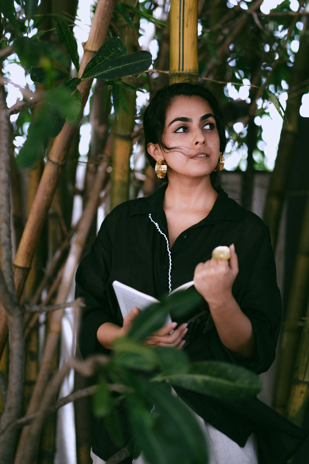 Woman in a black outfit holding a book among green plants wearing Japa Pushpa Hoops and a statement ring.