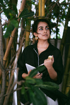 Woman in a black outfit holding a book among green plants wearing Japa Pushpa Hoops and a statement ring.