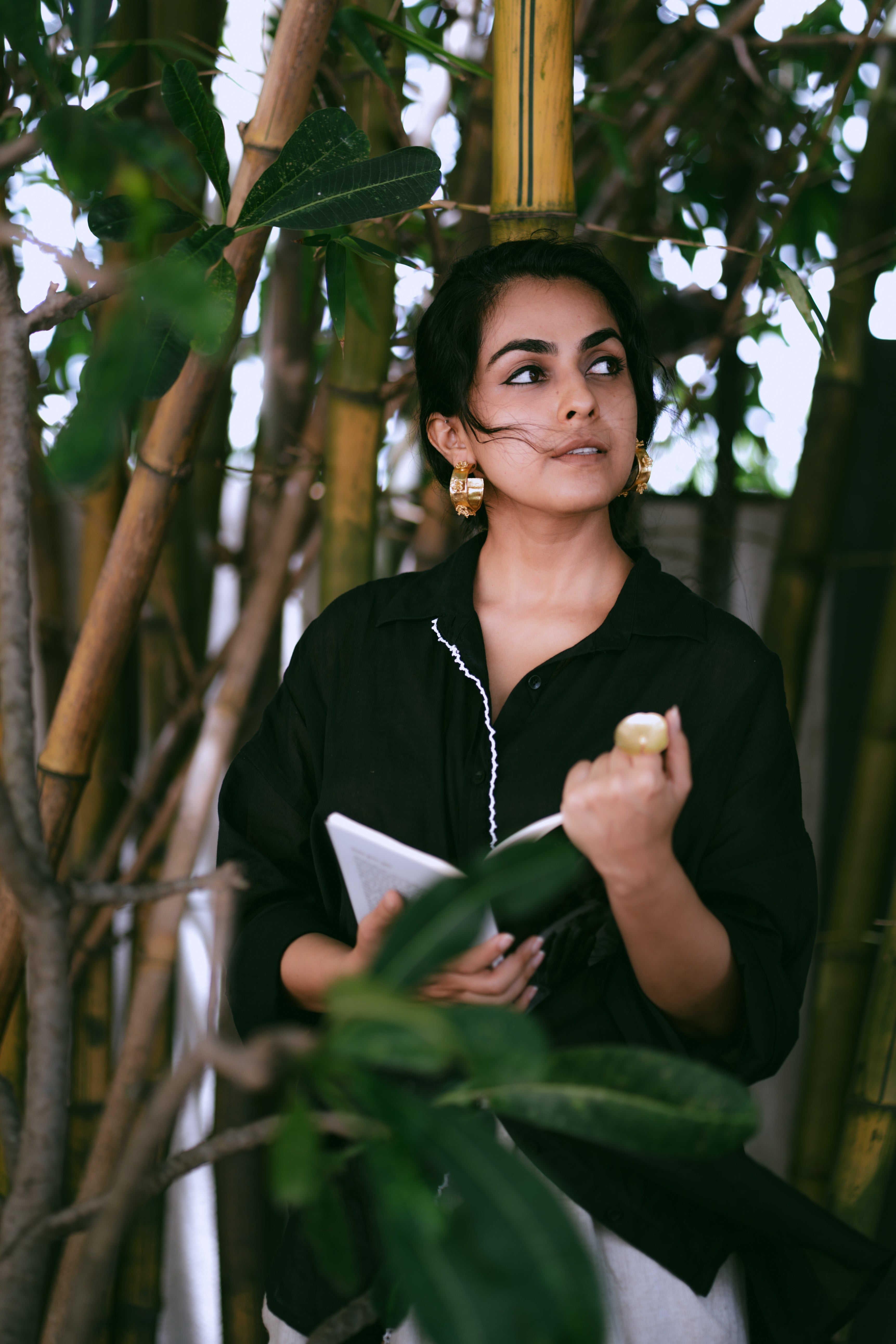 Woman in a black outfit holding a book among green plants wearing Japa Pushpa Hoops and a statement ring.