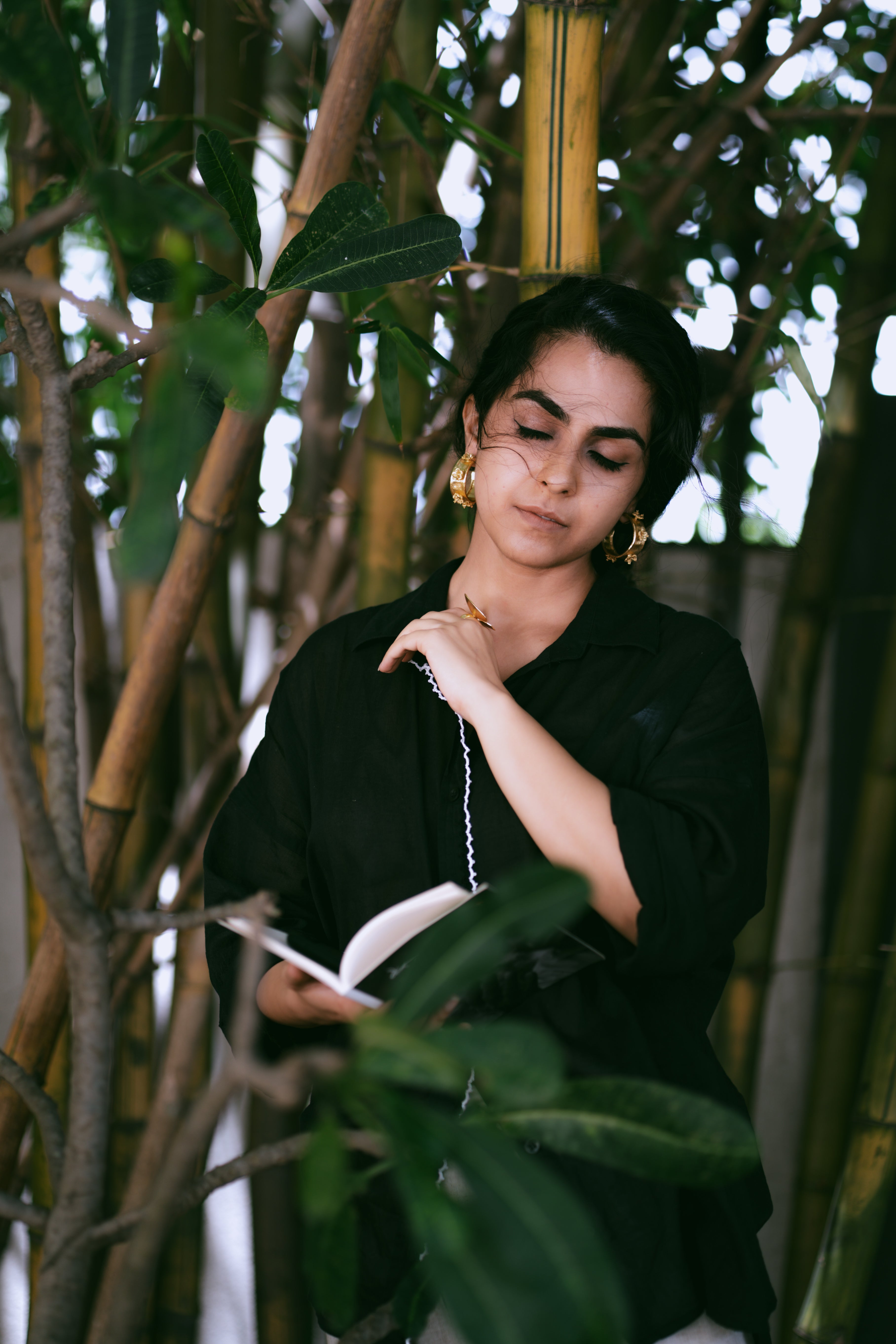 Woman in a black outfit holding a book among green plants wearing Japa Pushpa Hoops