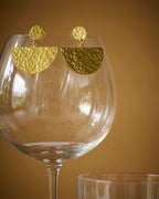 Gold plated round with half circle hammered drop earrings displayed on a wine glass against a mustard background