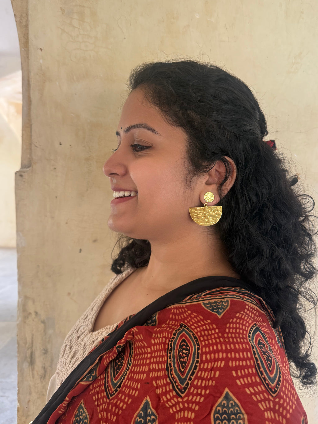 Woman wearing gold plated earrings and a red patterned dupatta, standing against a beige wall.