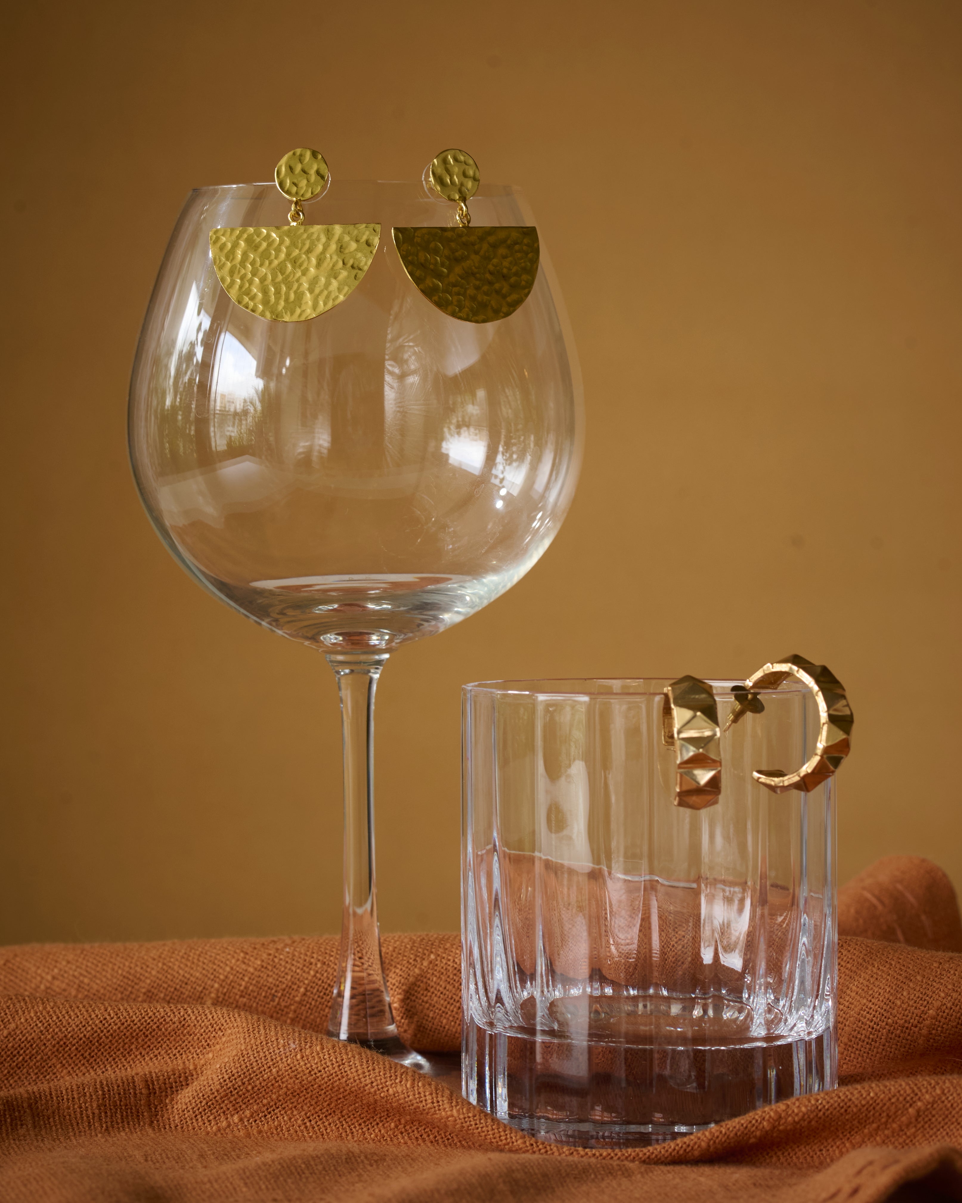 Gold earrings displayed on a glass with a mustard background