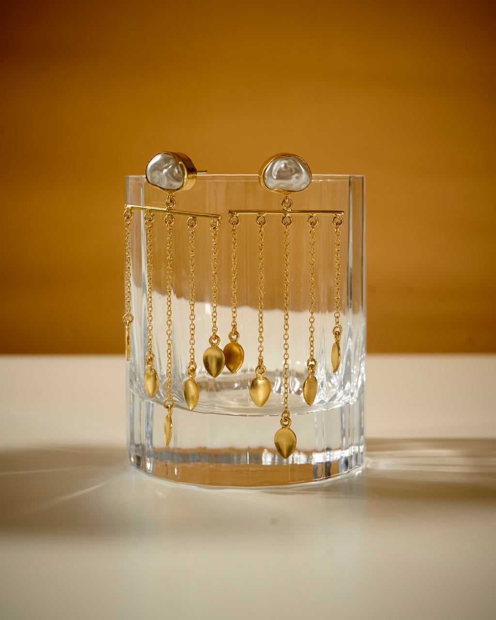 Clear glass holder with baroque pearl and gold color dangling earrings on a mustard background