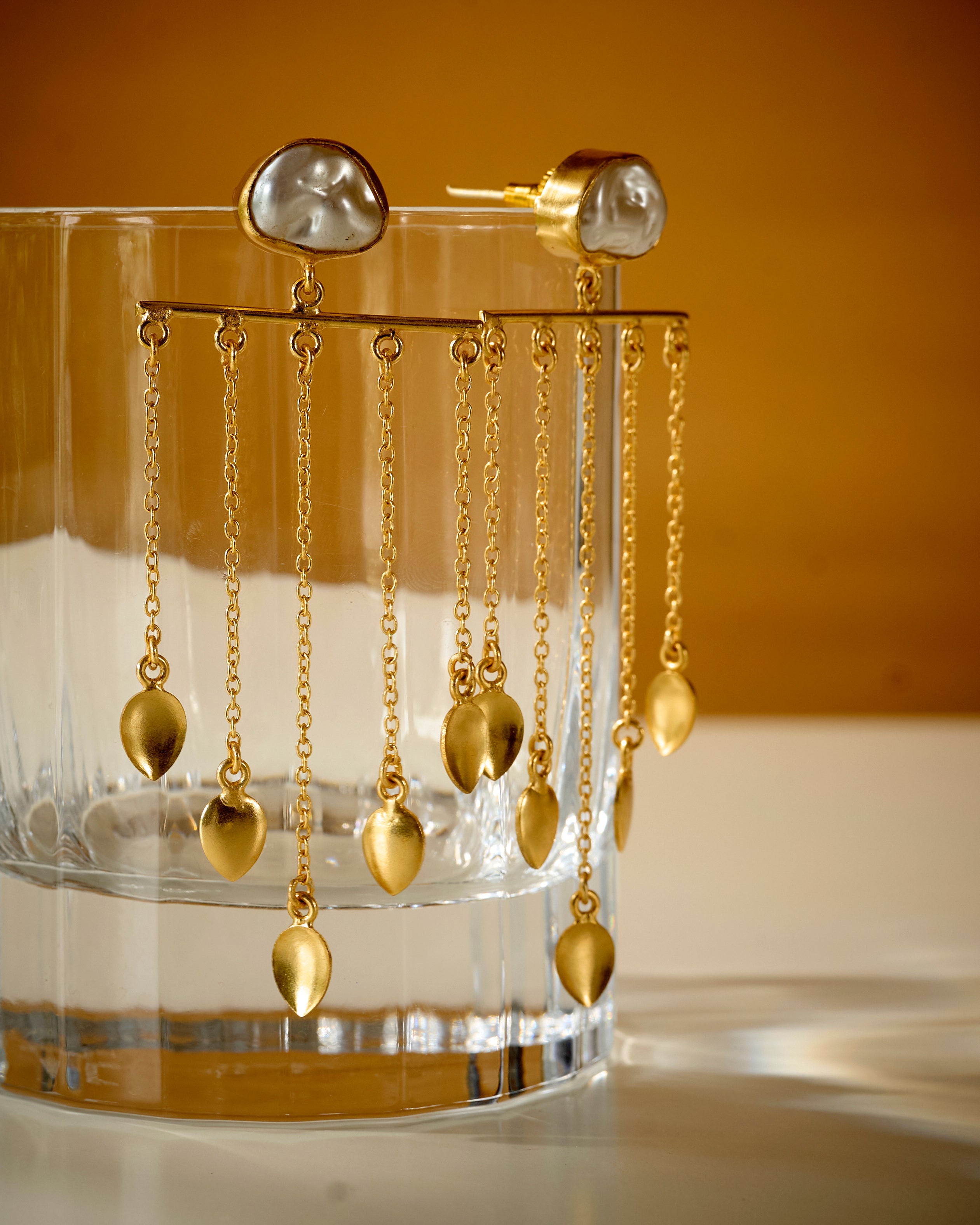 Close up of Clear glass holder with baroque pearl and gold color dangling earrings on a mustard background