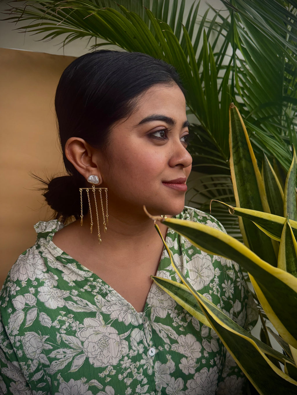 Woman wearing a green and white floral top with earrings, standing in front of plants.