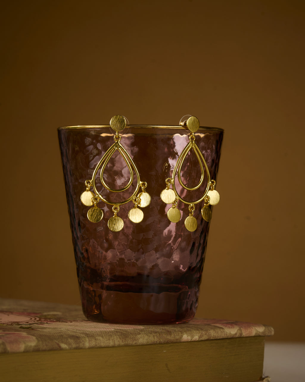 Gold earrings with dangling elements on a reflective glass against a mustard color background