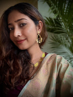 Woman wearing a saree with gold plated dangling earrings against a green leafy background.