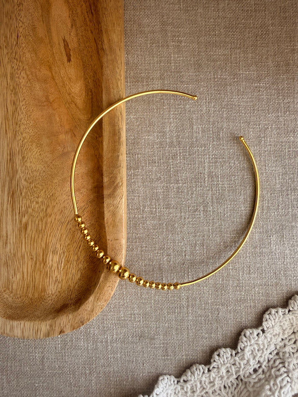 Kanak choker flatlay showing open wire design and gold bead cluster, placed across a wooden bowl and grey linen fabric with white crochet trim