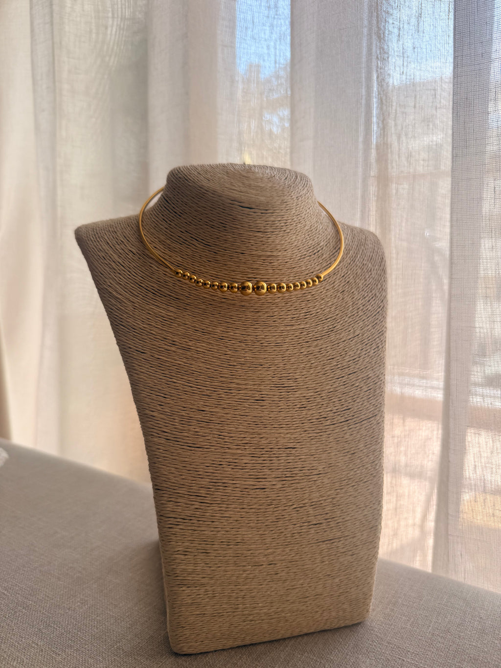 Gold necklace on a brown mannequin bust against a sheer curtain background