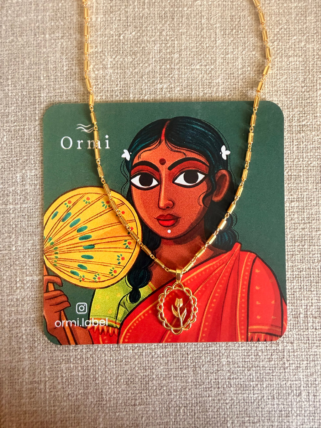Lily Necklace draped over an Ormi branded packaging card featuring a traditional Indian woman illustration in a red saree, placed on natural linen texture.