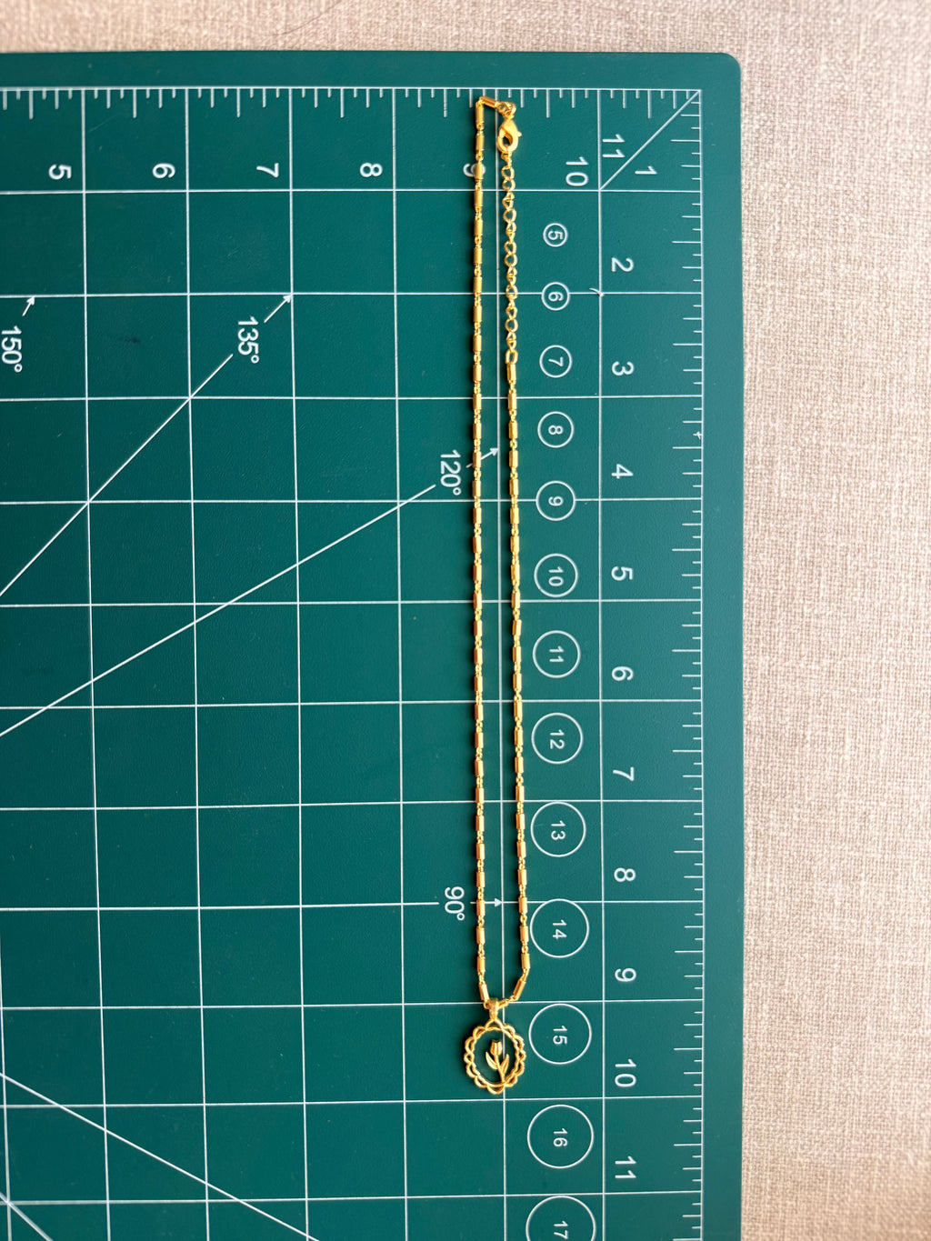 Full length flat lay of the Lily Necklace on a green cutting mat with ruler, showing the complete barrel-link chain length and oval lily pendant at approximately 10 inches.