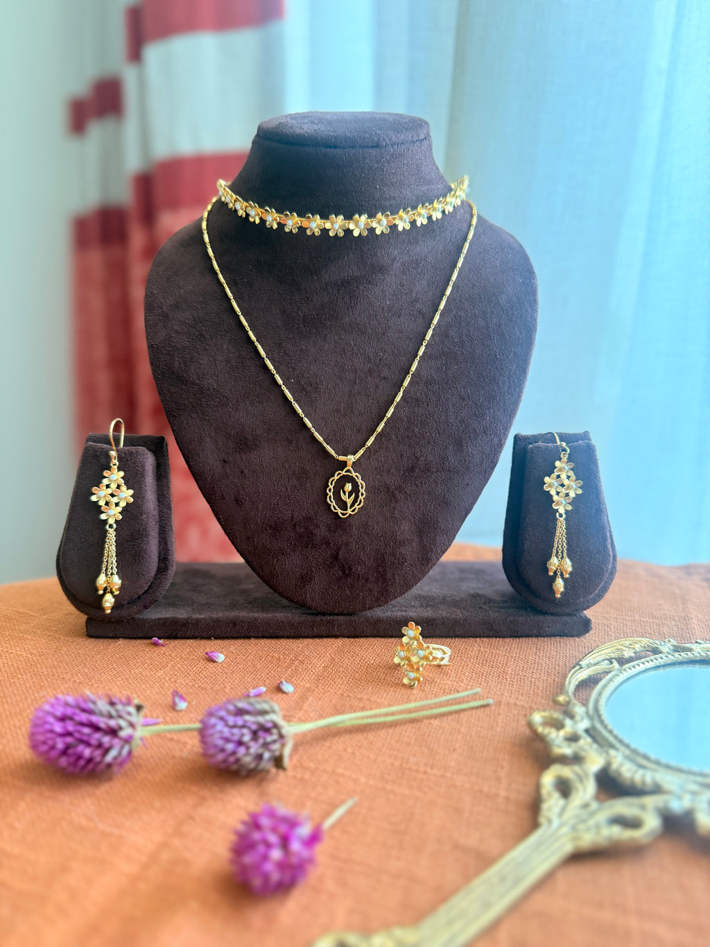 Full Motiya Capsule Collection displayed together — Motiya Choker and Lily Necklace layered on a dark velvet bust, Motiya Danglers on small velvet stands on either side, Motiya Lily Ring in the foreground, surrounded by dried purple globe amaranth flowers and a vintage brass mirror on terracotta linen.