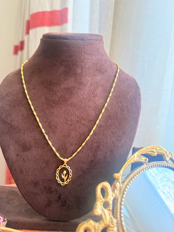 Gold plated brass Lily Necklace with oval scalloped pendant holding a lily bud, on a barrel-link chain, displayed on a dark velvet bust with a vintage brass mirror partially visible in soft natural light.