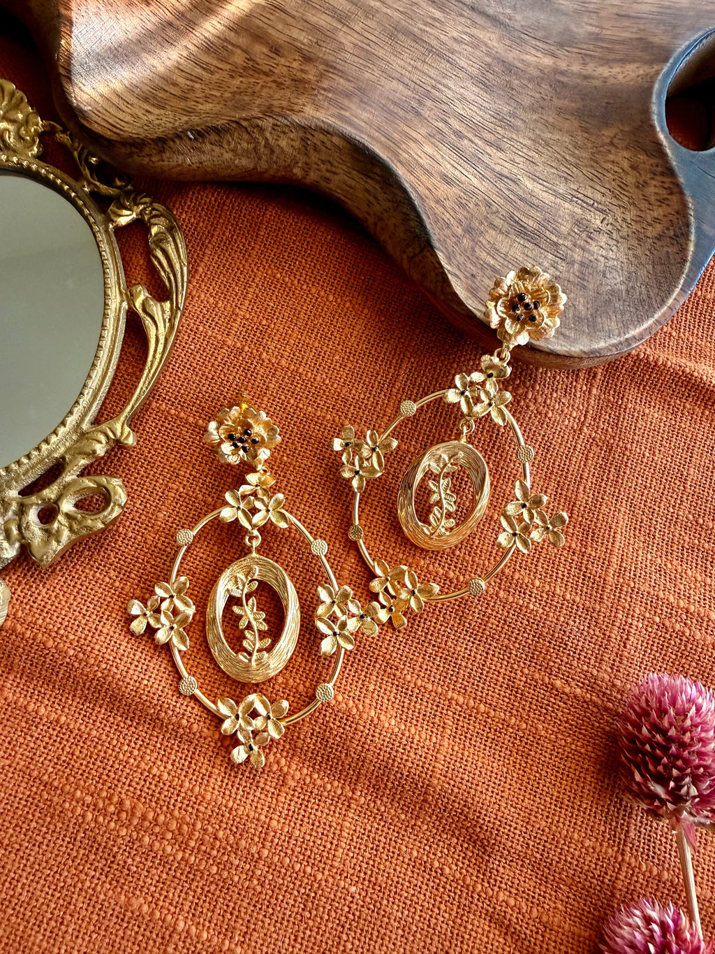 Pair of gold Malika Danglers with a small flower stud and an oval frame of tiny blossoms around a leafy sprig, laid on rust linen beside a wooden tray and brass mirror