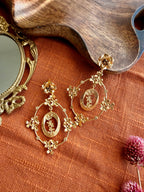Pair of gold Malika Danglers with a small flower stud and an oval frame of tiny blossoms around a leafy sprig, laid on rust linen beside a wooden tray and brass mirror