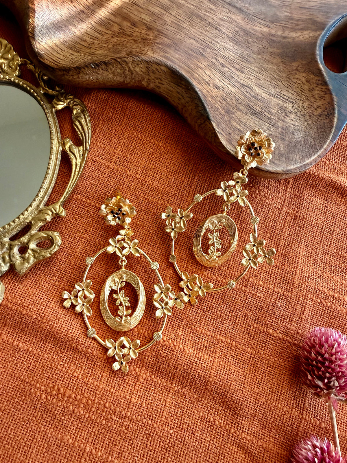 Pair of gold Malika Danglers with a small flower stud and an oval frame of tiny blossoms around a leafy sprig, laid on rust linen beside a wooden tray and brass mirror
