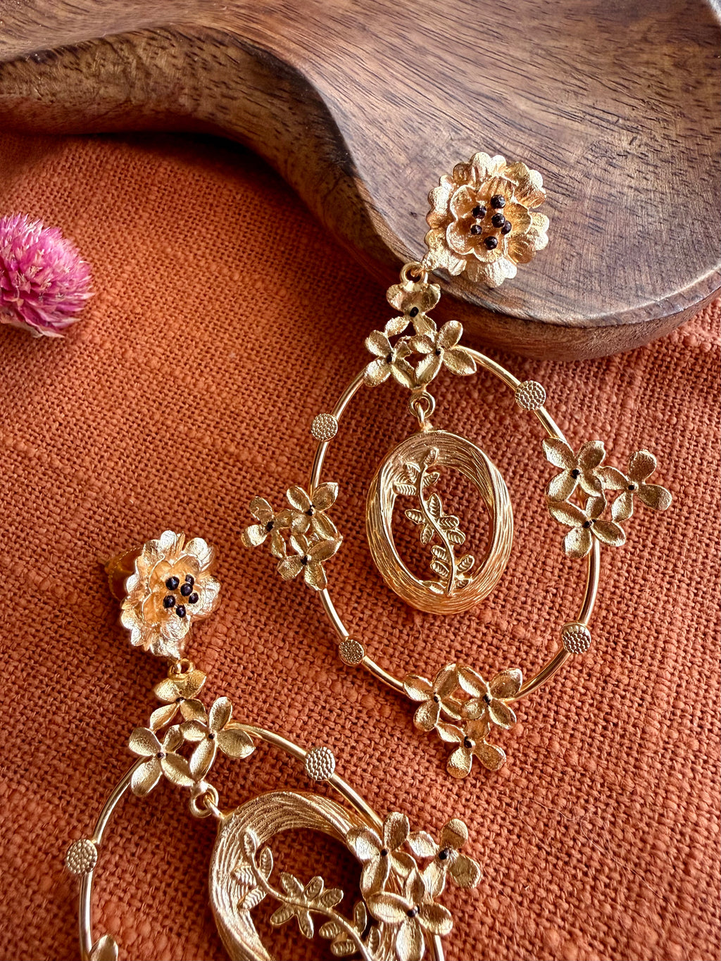 Close-up of Malika Danglers on rust-orange linen, showing the flower stud, tiny blossoms and the textured oval centre with a leafy sprig