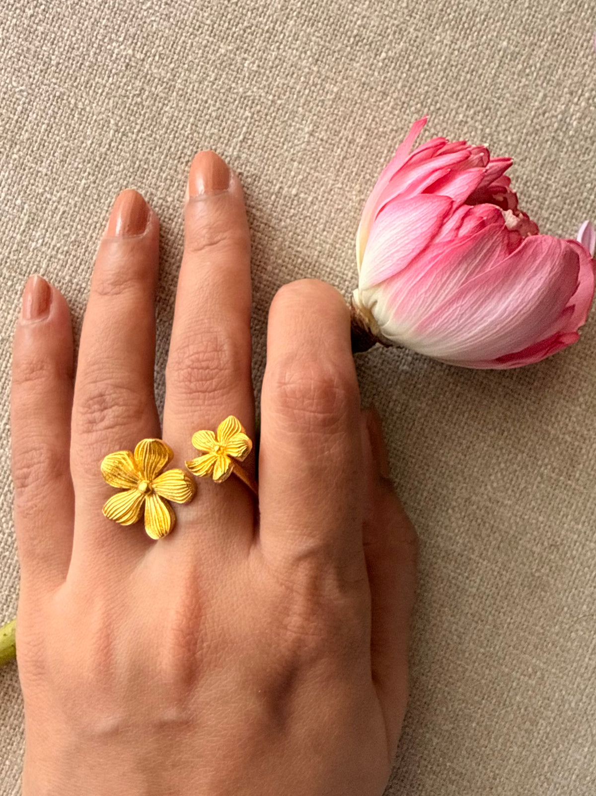 Model's hand showing the dual-flower open-wrap design of the gold Manjari Ring styled next to a pink lotus.