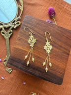Gold plated brass Motiya Danglers with jasmine flower cluster and pearl centres, three hanging chains with ribbed gold beads, displayed on a dark wooden box with a vintage brass mirror and dried purple flowers on terracotta color fabric.