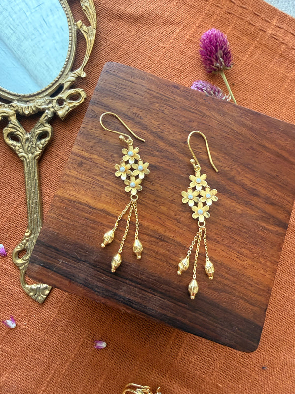 Gold plated brass Motiya Danglers with jasmine flower cluster and pearl centres, three hanging chains with ribbed gold beads, displayed on a dark wooden box with a vintage brass mirror and dried purple flowers on terracotta color fabric.