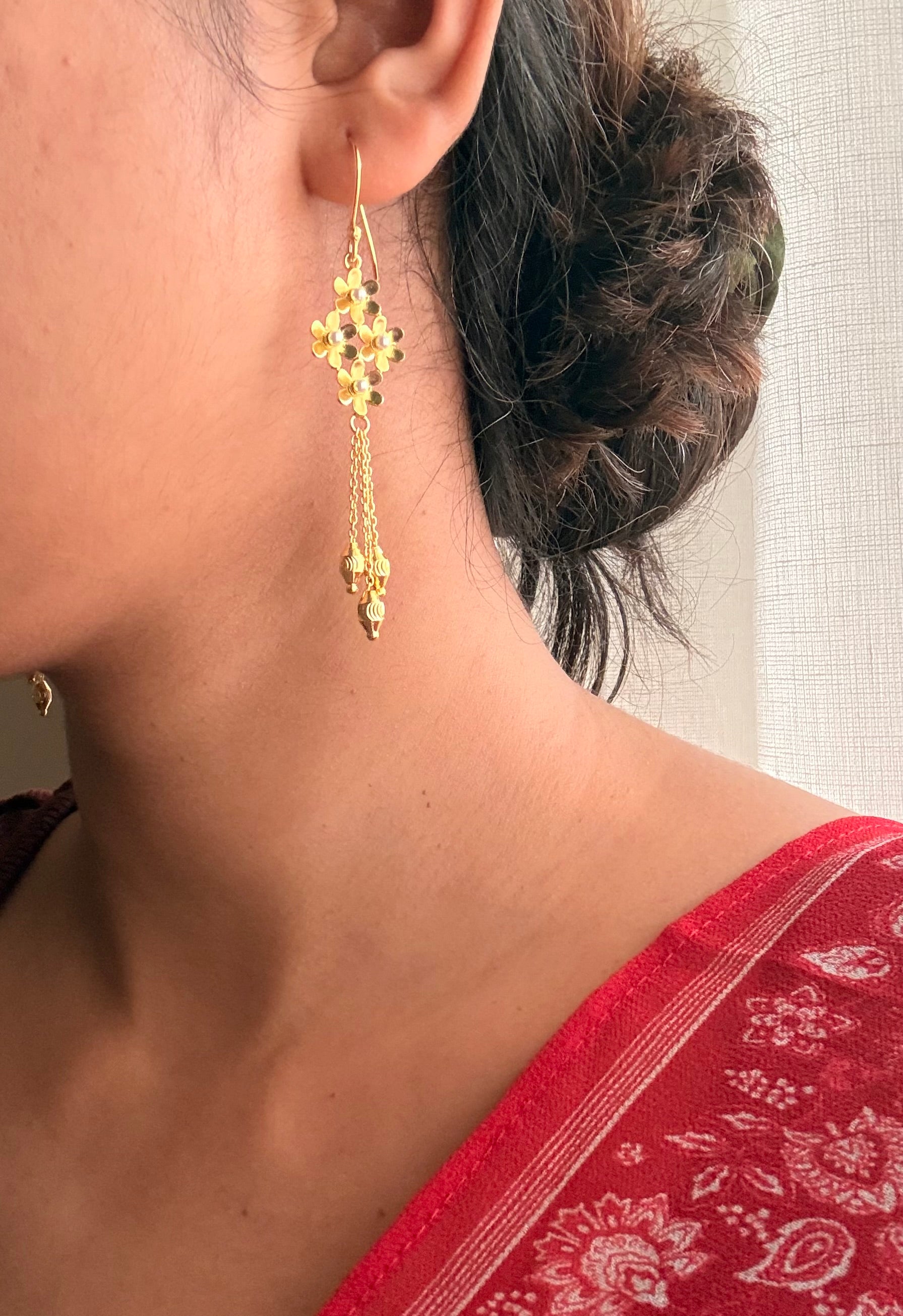 Close-up side profile of Motiya Danglers on the ear, showing the flower cluster and gently swaying chains against a red floral saree border, with soft diffused window light.