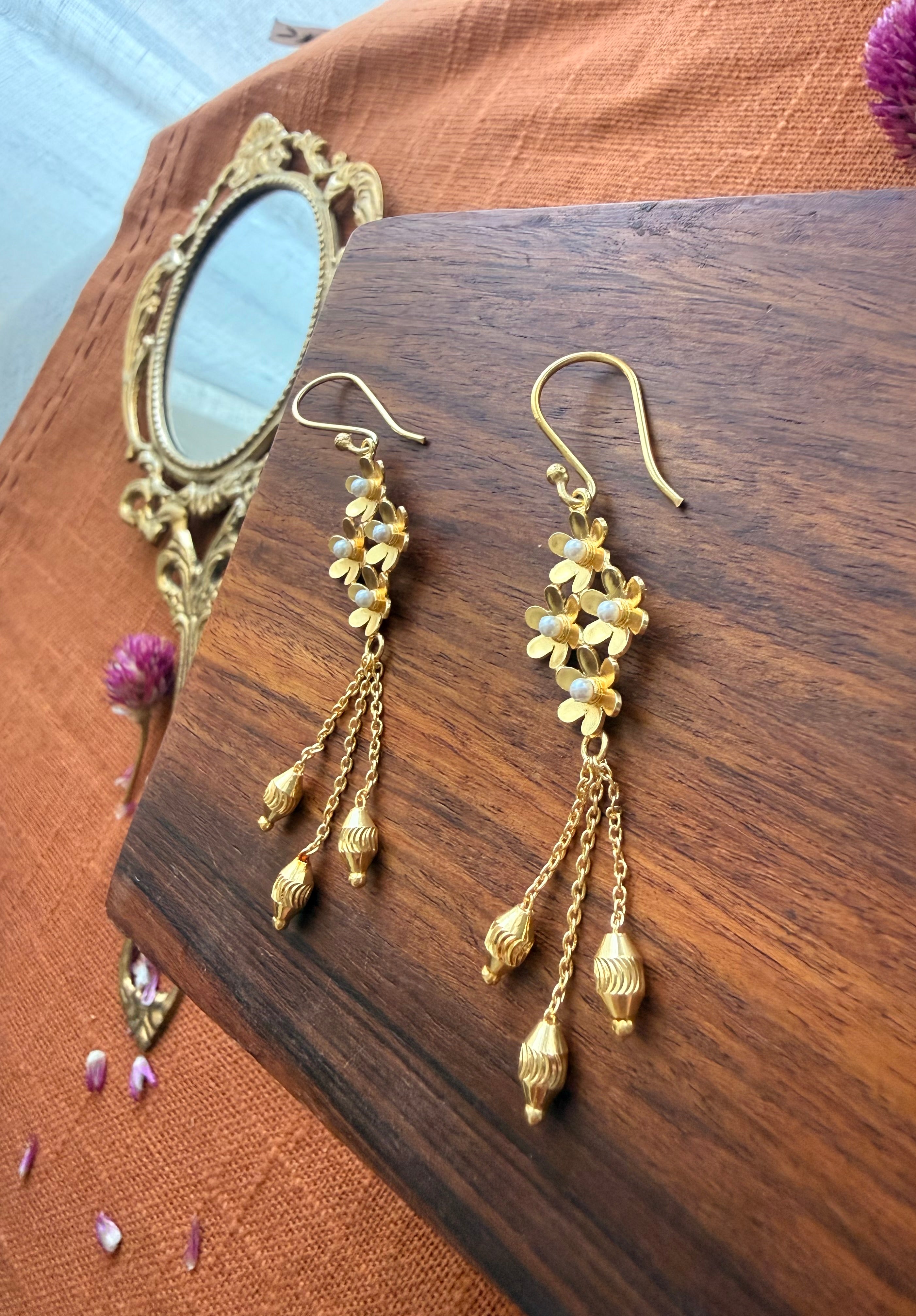 Flat lay of Motiya Danglers showing pearl-centred jasmine flowers and three-chain tassel with ribbed gold beads, displayed on dark walnut wood with a vintage oval brass mirror and scattered dried purple petals on rust linen.