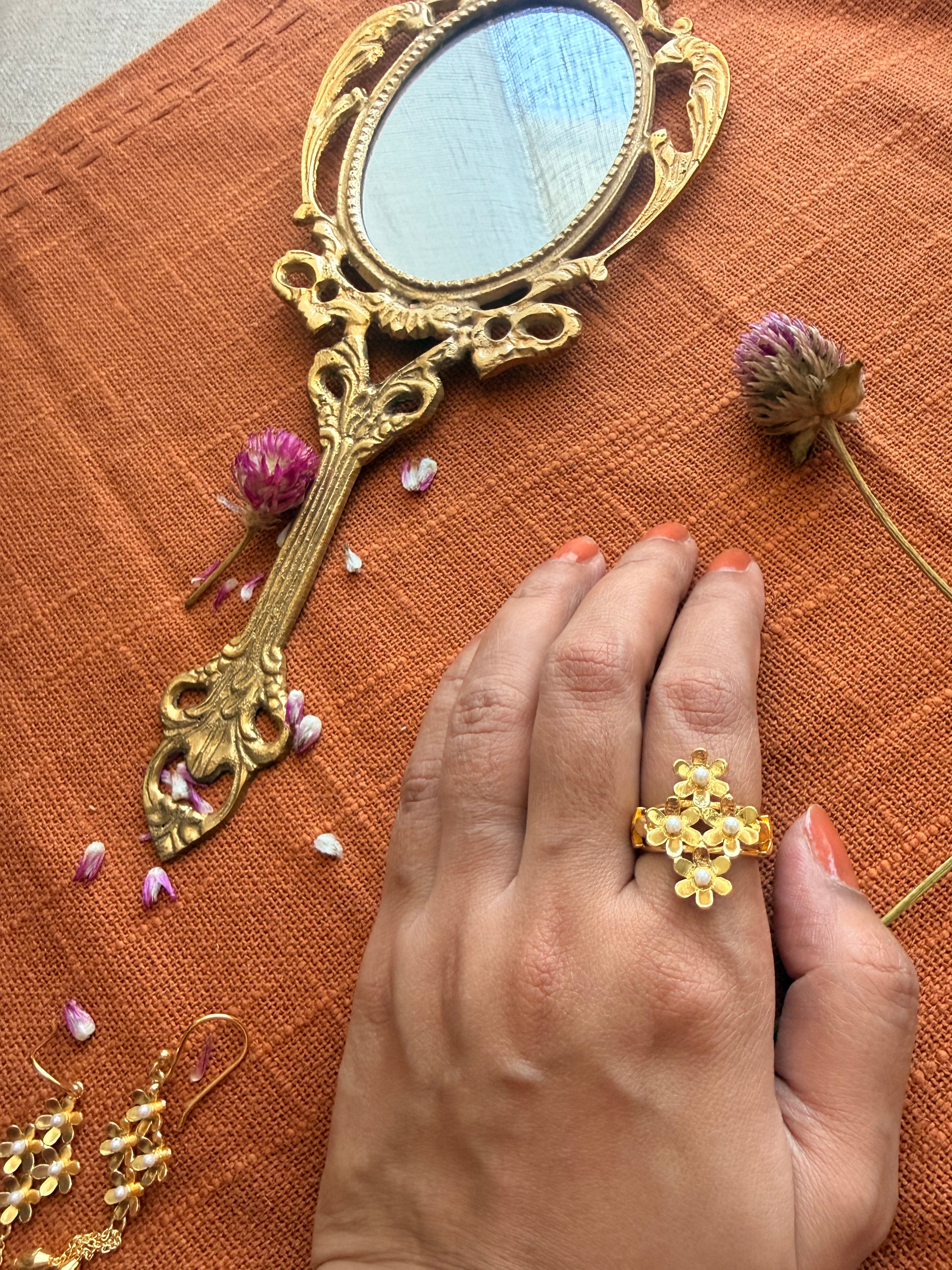 Motiya Lily Ring with jasmine flower cluster and pearl centres worn on the finger, photographed flat lay style top view on terracotta linen with a vintage brass hand mirror, scattered dried purple flowers.