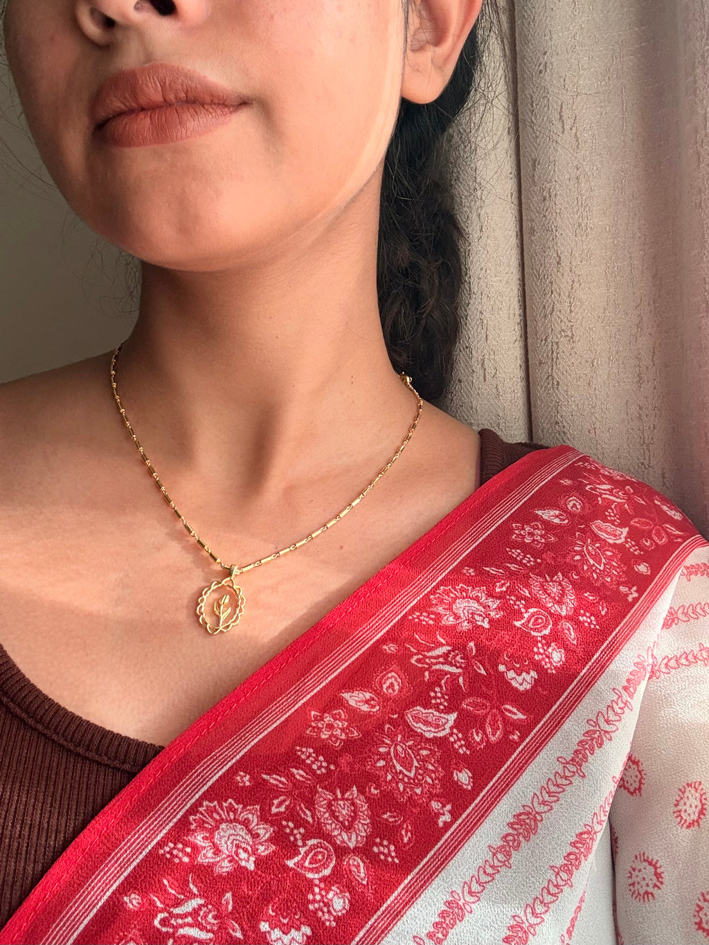 Lily Necklace with oval scalloped pendant and lily bud worn close to the collarbone, styled with a brown fitted top and red floral printed dupatta, photographed in warm side light against a neutral wall.